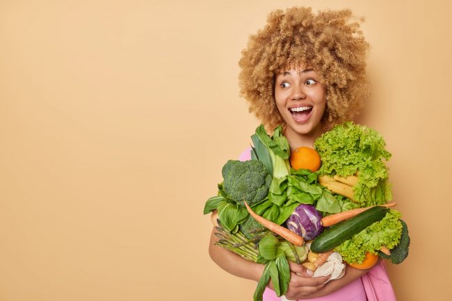 Horizontal shot of happy surprised woman reacts to something funny carries freshly picked vegetables from garden keeps to clean eating prefers vegetarian food poses indoor empty space for text